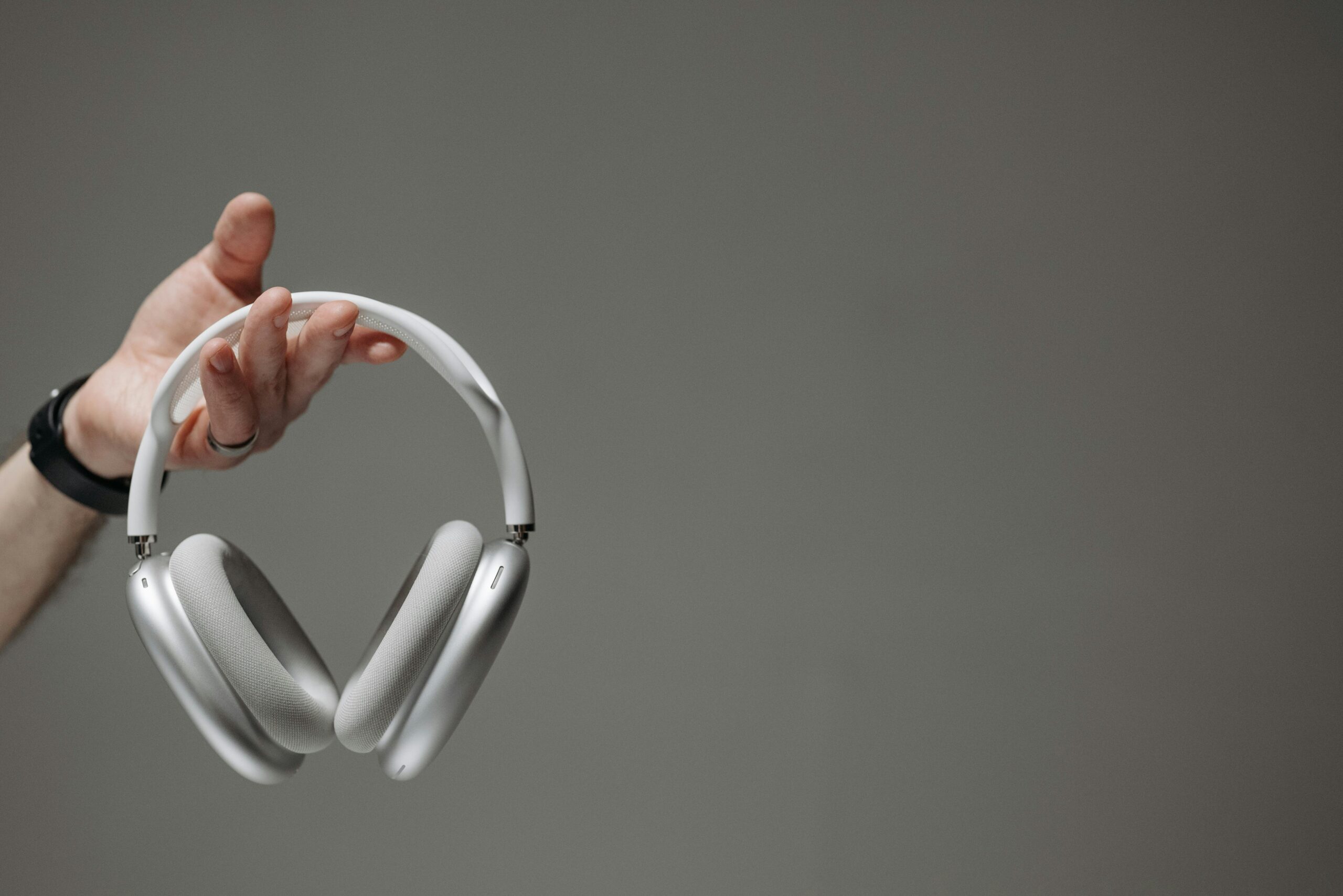 White wireless headphones held by a hand against a neutral gray background.